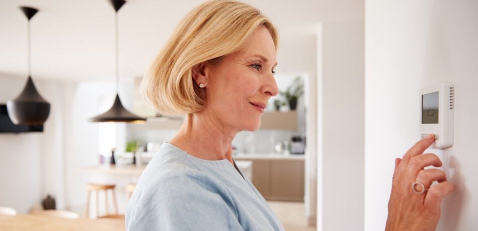 A blonde, older woman touching the thermostat on the wall in her home.