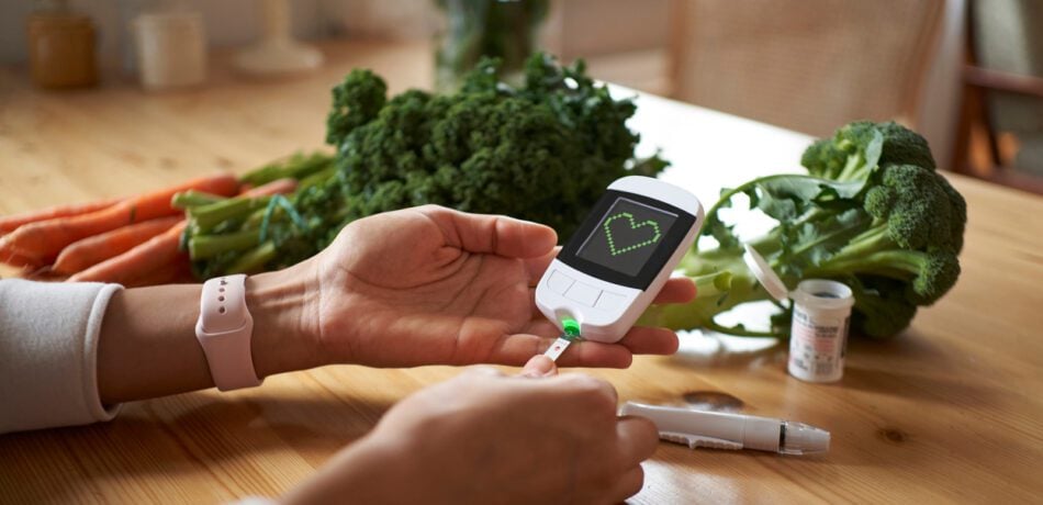 A person using a blood glucose monitor with fresh vegetables in front of them on the table.