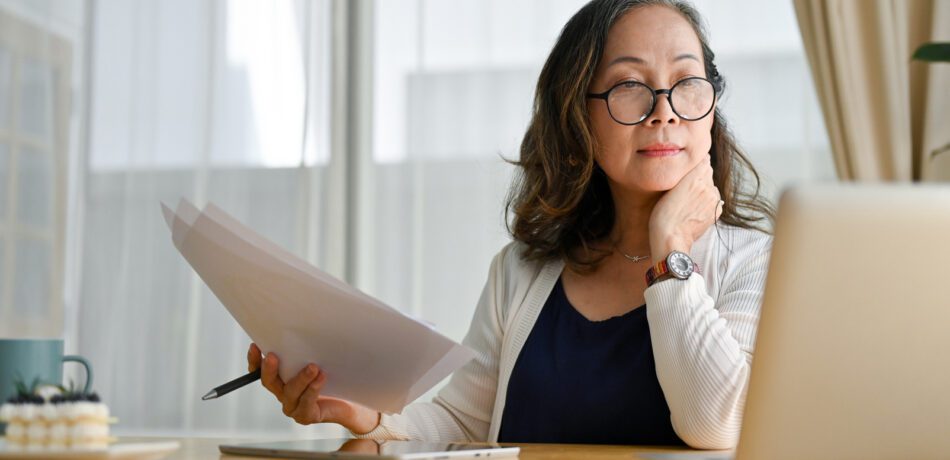 A woman wearing glasses working on a computer while holding papers.