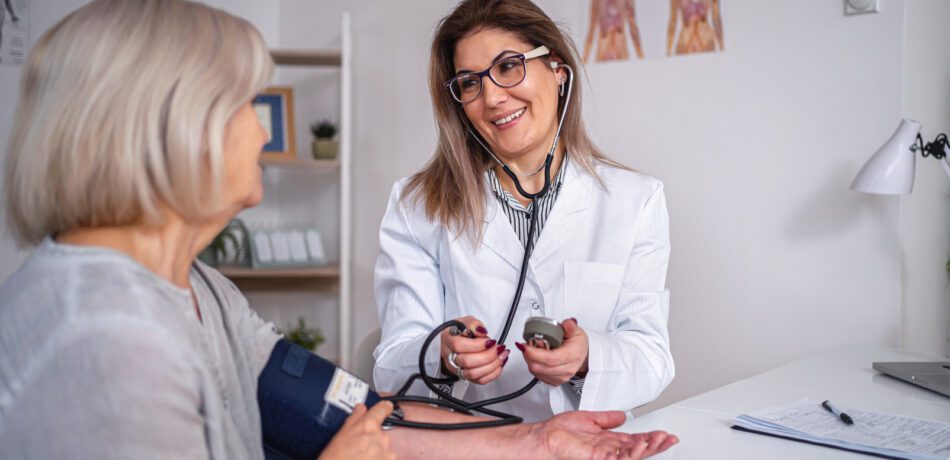 A woman medical professional taking another woman's blood pressure as they chat and smile while seated.