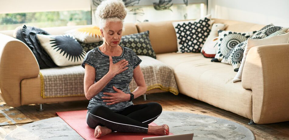 A woman sitting on a yoga mat in front of her couch meditating with her hands on her chest and stomach while her eyes are closed.