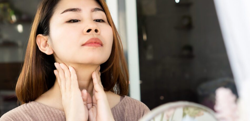 Asian woman looking in mirror while feeling her neck for a self exam.