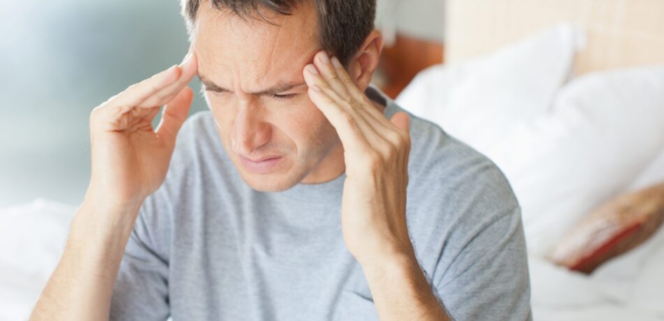 Man with gray t-shirt sitting on bed looking pained and rubbing his temples.