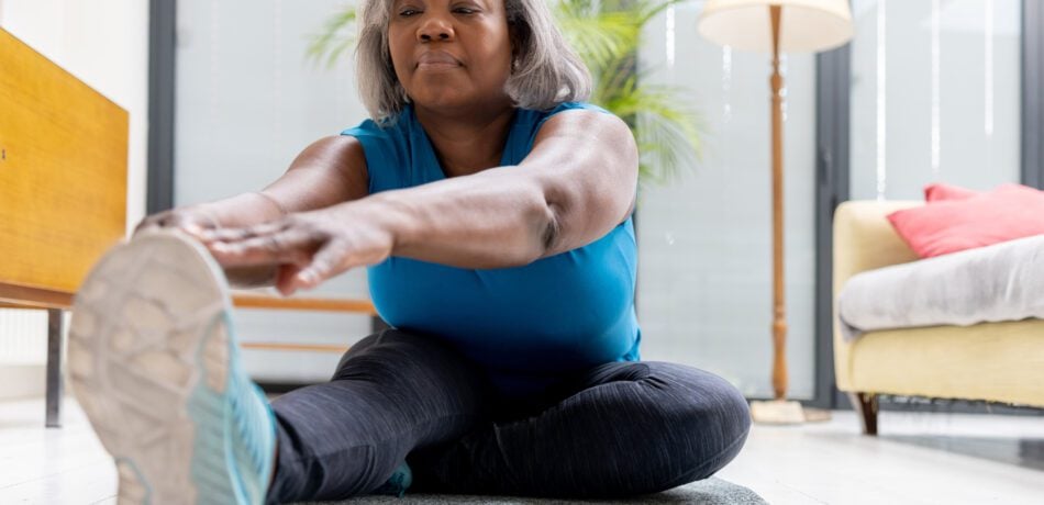 Mature woman stretching her arms and legs while sitting on a mat in her living room.