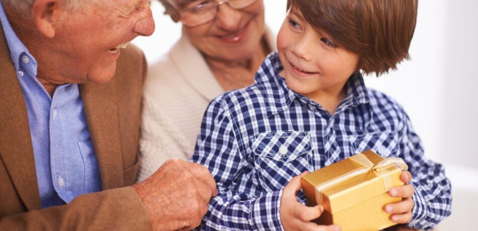 A young boy sitting on his smiling grandmother's lap while holding a wrapped gift as his grandfather smiles at him.