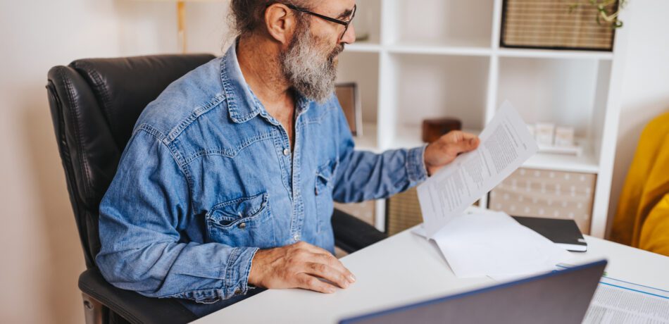 Mature man in blue shirt going through paperwork while working on a laptop at home.