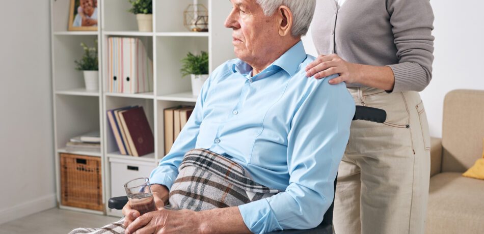 Sad looking older man sitting in a wheelchair with a caregiver standing behind him.