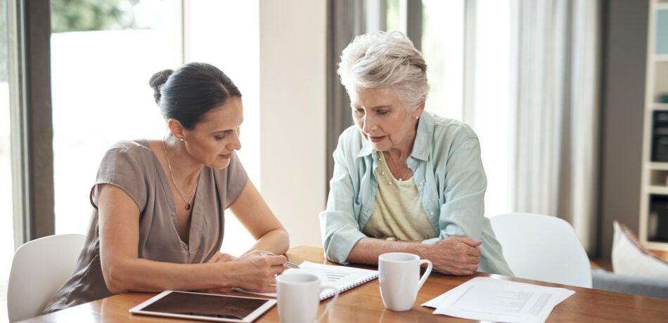 Adult woman and her senior mother sit at a table with coffee cups looking down at a notebook together.