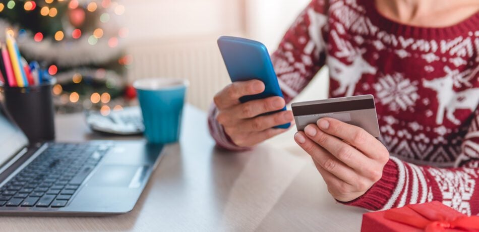 Woman wearing red sweater at table holding a credit card and her phone with a laptop in front of her.