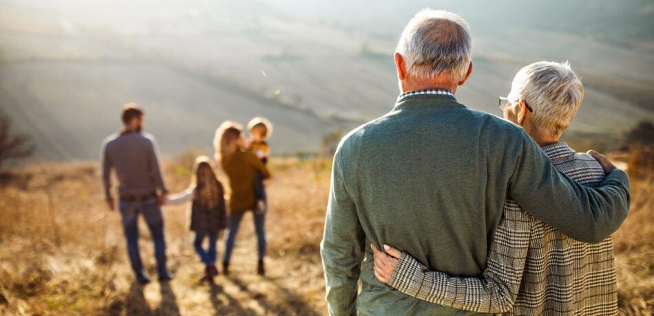 Back view of grandparents arm in arm while looking at their family ahead of them in a field with hills in the background.