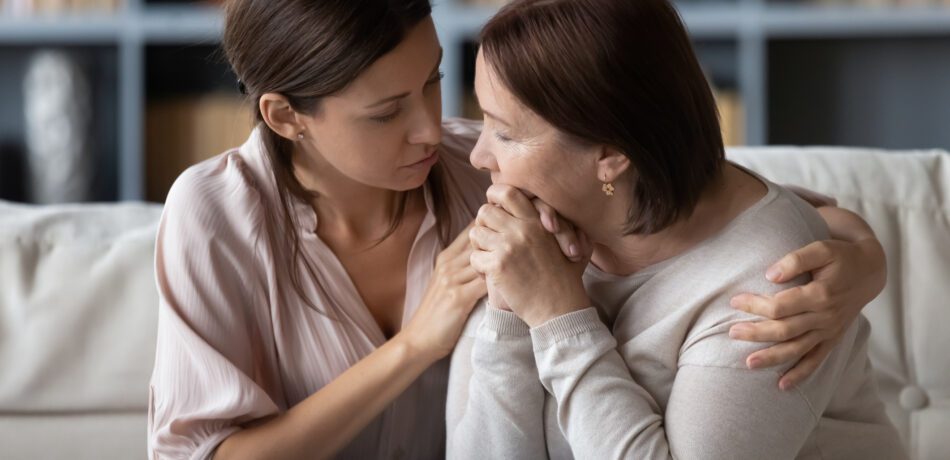 Adult daughter with arm around her grieving mom while both sit next to each other on a couch.