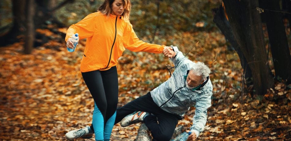 Senior couple on a path in a forest with the man stumbling over a tree limp while the woman holds his hand to help.