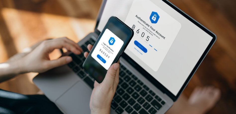 Cropped hand of woman using a phone in front of a laptop to verify her login.