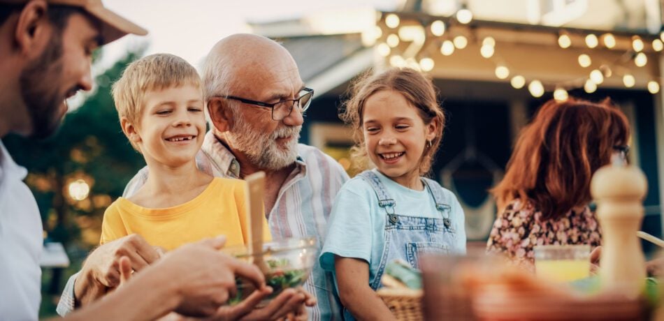 Older grandfather sitting at table outside with smiling grandkids on his lap and other family members around them.