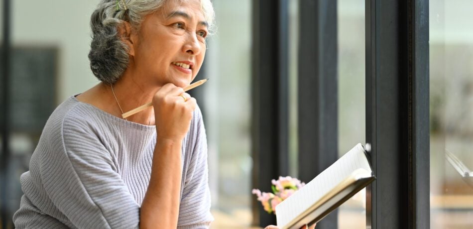 Mature woman smiling holding a journal and pencil as she looks out a window.