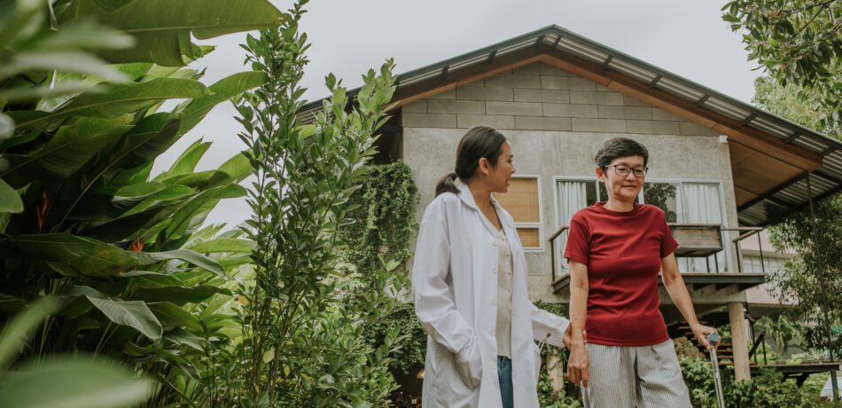 Female doctor holds woman’s hand as she walks with a cane outside.