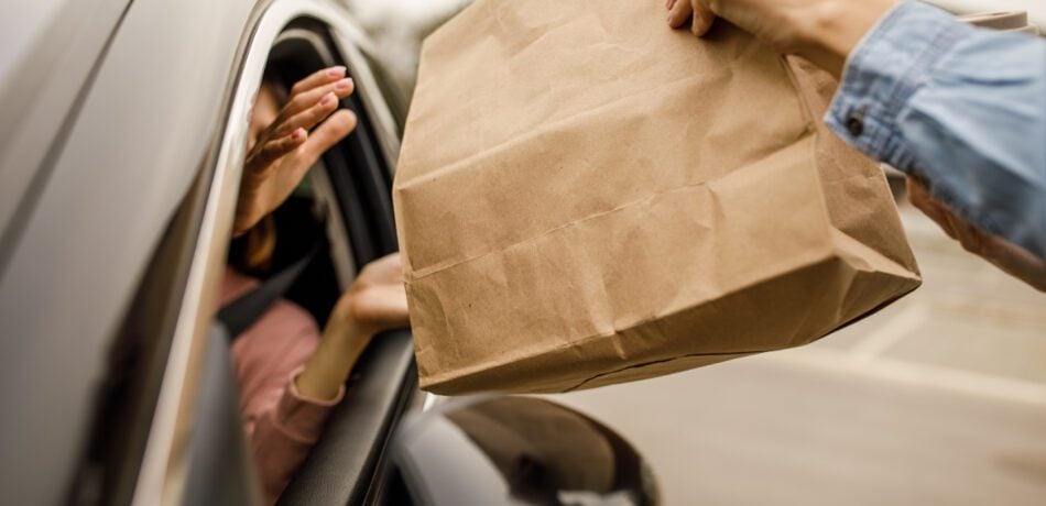 Close up shot of unrecognizable customer sitting in her car, in driver's seat, receiving a coffee and takeaway food order from a service person at the drive through.