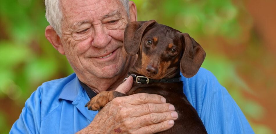 Older man smiling while holding a brown Dachshund dog.