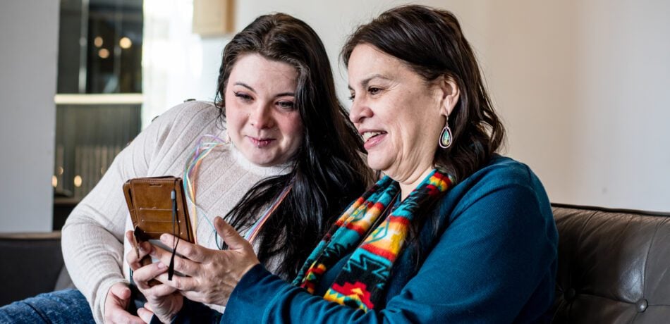 Mother and adult daughter sit on couch looking at phone and smiling.