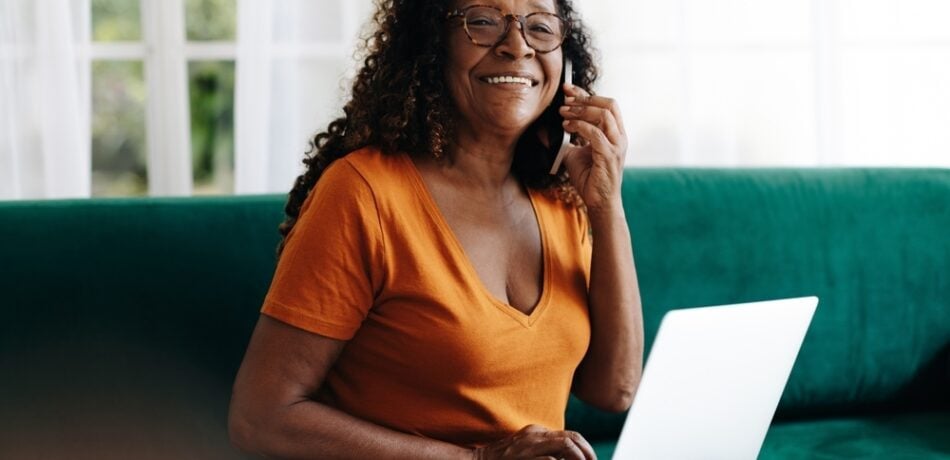 Mature woman talks on the phone while working on her laptop from the couch.