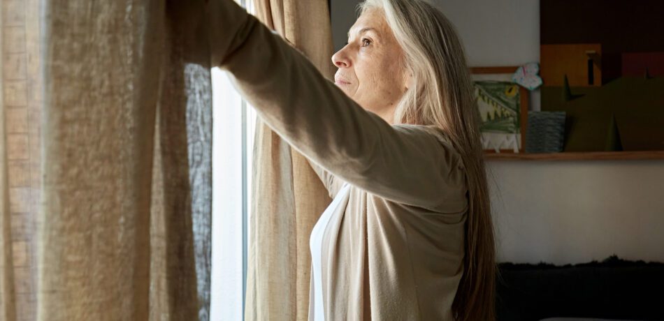 Older woman opens the curtains in her home.