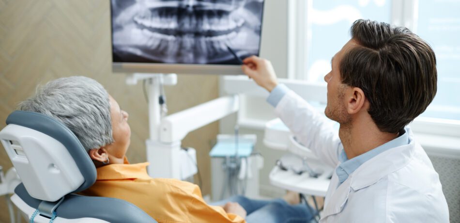 Back view of male dentist pointing at tooth X-ray image on screen during a dental appointment with a patient in the chair looking on.