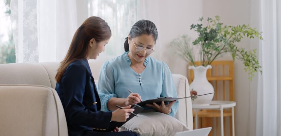 An older woman signs papers with her adult daughter by her side also holding a pen.