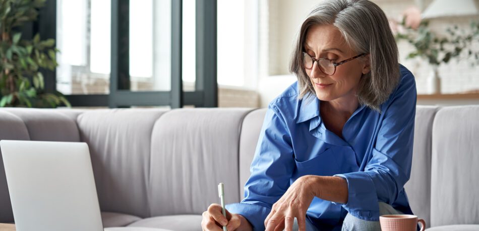 A woman works on a laptop and takes notes in a book.