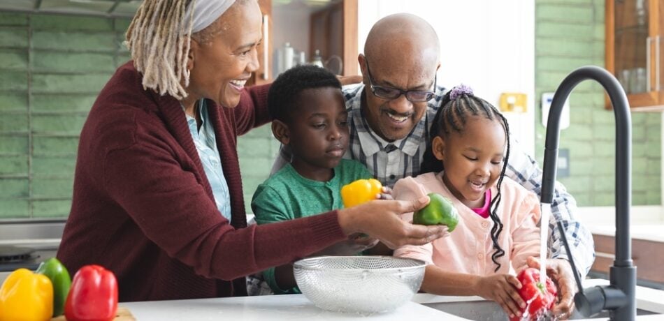 Grandparents and grandchildren wash vegetables in a kitchen sink.