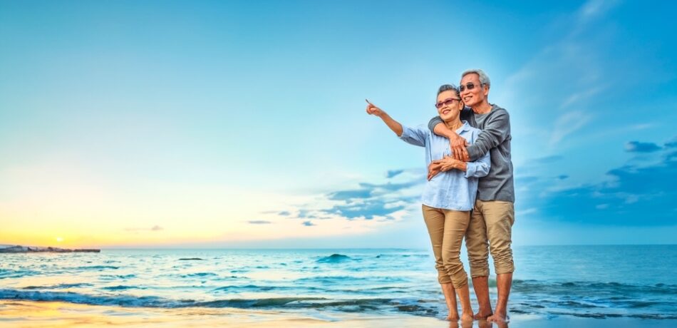 An older couple embraces on a beach while pointing into the distance.