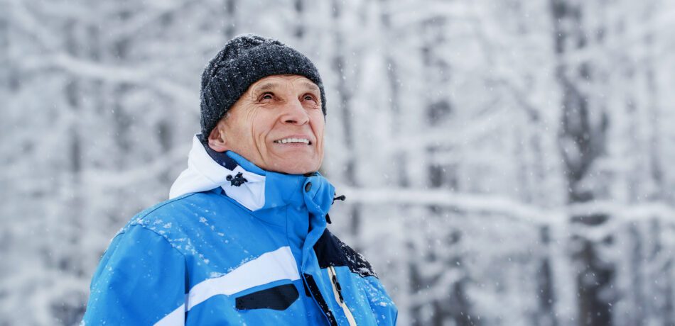 Older man smiling in blue winter coat and black hat as he walks through a snowy forest.