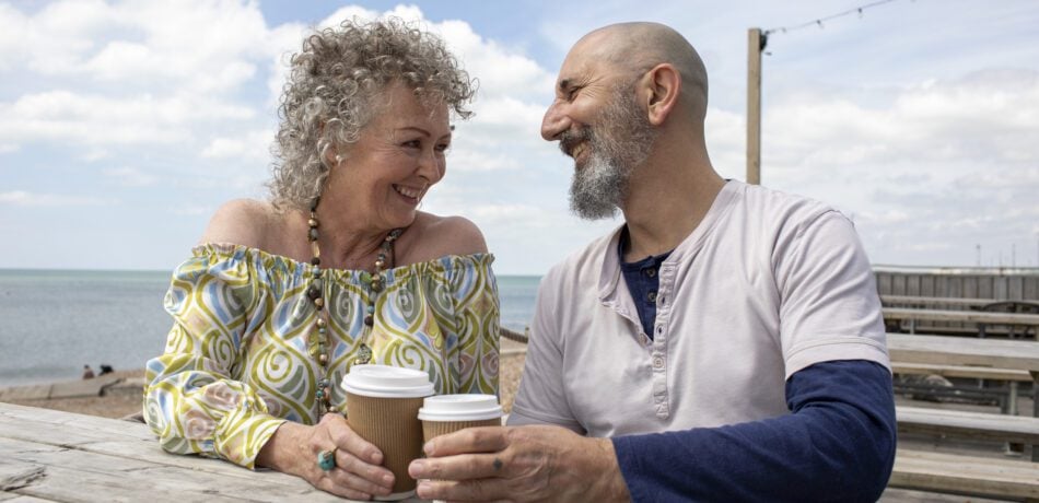 Mature couple enjoying coffee together by the beach at a picnic table.