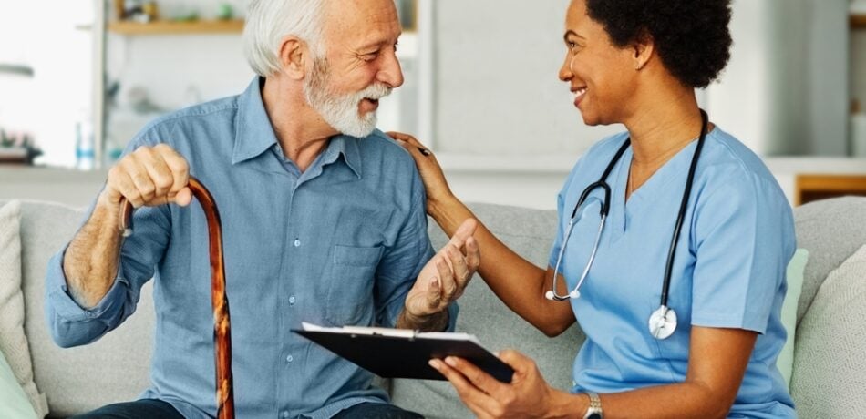 An older man sits with a nurse in his home.