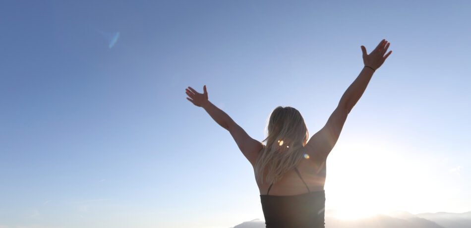 Back of a woman shown as she lifts her hands over her head looking at the sunrise with thankfulness.