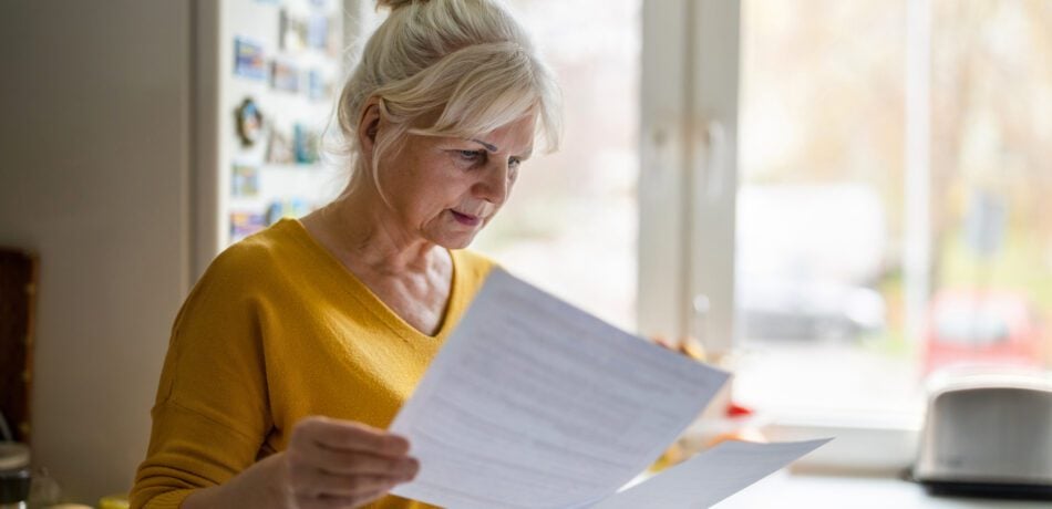 Older woman in her kitchen looking serious while reading two medical billing statements.