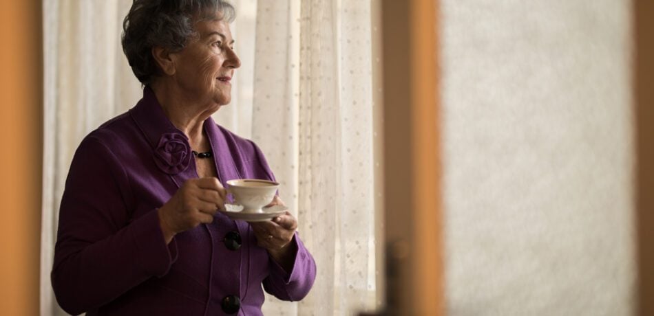 Senior woman drinking a cup of tea looking out of her window.