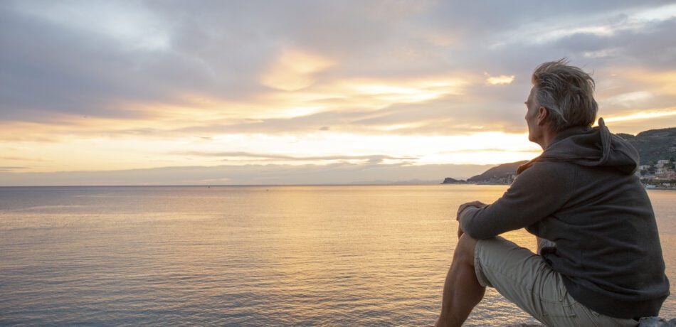 Man looks out to sea from coastal area at sunrise.