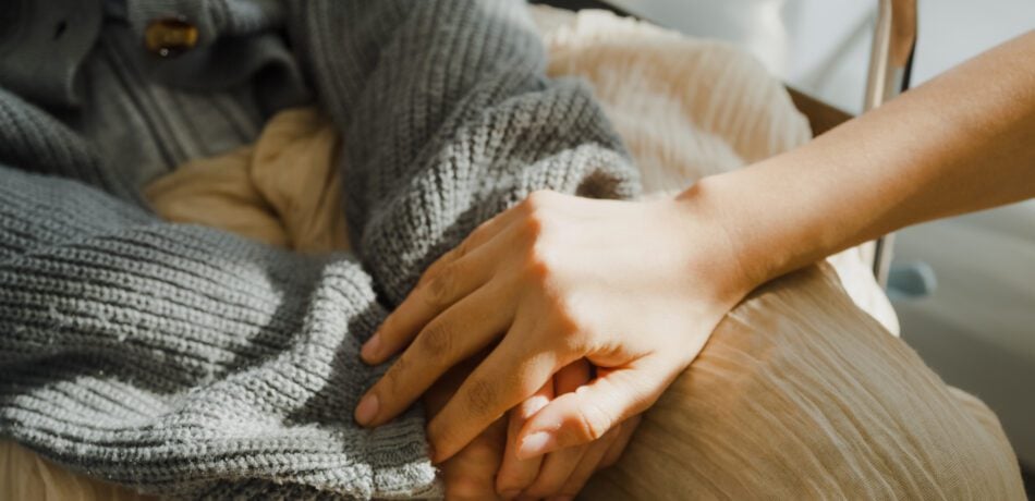 Closeup of young caregiver's hand on the hand of an older adult wearing a sweater and using a blanket.