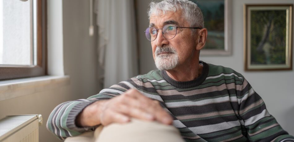 A senior man pensively sits on a sofa by the window looking outside.